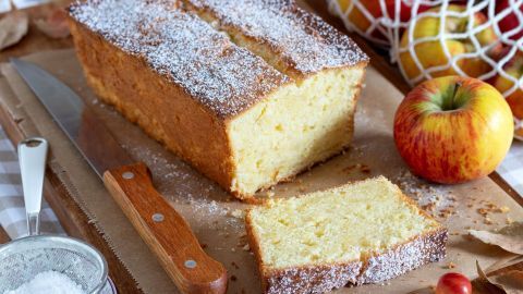 Ein saftiger Rührkuchen mit zuckrigem Überzug liegt auf einem Holzbrett, neben einem geschnittenen Stück und einem Apfel.
