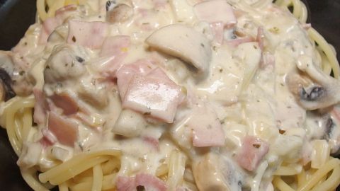 Nudeln in Sahnesoße liegen cremig auf einem Teller, umgeben von würzigen Schinkenstücken und frischen Champignons.