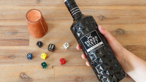 Eine Hand hält eine Flasche zu einem Trinkspiel, umgeben von Würfeln und einem Becher auf einem Holztisch.