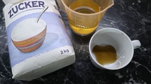 Ein Sack Zucker liegt neben einem Messbecher mit Karamelsirup und einer Tasse. Der Sirup glänzt goldbraun.