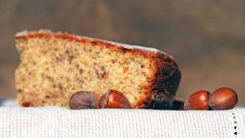 Ein Nusskuchen liegt auf einem Stofftuch, umgeben von Haselnüssen, im Hintergrund unscharf.