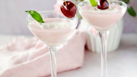 Zwei elegante Cocktails aus Kirsch-Joghurt-Likör stehen auf einem Tisch, umgeben von zarten Blumen und einem pinken Tuch.