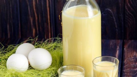 Eine Flasche Eierlikör steht neben zwei Gläsern mit dem cremigen Getränk und frischen Eiern auf einem rustikalen Holzuntergrund.