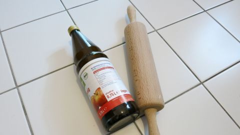 Kein Nudelholz zur Hand und du möchtest Pizza oder Plätzchen backen? Nimm einfach eine Flasche als Ersatz. Eine Holzrolle und eine Flasche liegen auf einem weißen Fliesenboden, bereit für einfache Küchenarbeiten ohne Nudelholz.