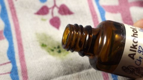 Mit Alkohol lassen sich Spinat- oder Grasflecken effektiv entfernen. Einfach auf den Fleck auftragen und der Natur beim Verschwinden zusehen. Im Bild wird die Methode an einem befleckten Tuch demonstriert. Ein braunes Fläschchen mit der Aufschrift "Alkohol" wird über ein Tuch mit grünen Spinatflecken gehalten.