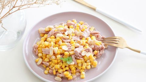 Auf einem rosa Teller liegt ein Salat aus Mais und Thunfisch, garniert mit einer Kräuterblatte und einer Gabel daneben.