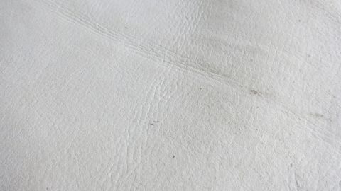 Extrem verschmutzte weiße Kunstlederpolster mit White Care Waschmittel wieder sauber bekommen. Ein glattes Stück weißes Kunstleder liegt flach, zeigt eine leichte Textur und kleine Unreinheiten, bereit zum Reinigen.