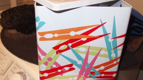 Eine bunte Box mit Wäscheklammern steht auf einer Waschmaschine, umgeben von Wäsche und Notizen zur Wäschepflege.