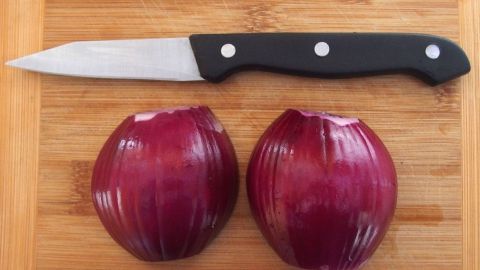 Zwiebeln gegen Wespenstiche - das Jucken lässt nach und es entsteht fast keine Schwellung. Rote Zwiebeln liegen halbiert auf einem Holzbrett, daneben ein scharfes Messer zum Schneiden.