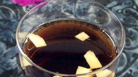 Ein Glas Cola mit Ingwerstückchen steht auf einem gemusterten Tisch, umgeben von lila blühenden Pflanzen.
