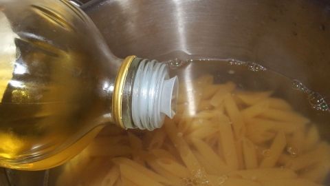 Eine Flasche Öl gießt Öl ins kochende Nudelwasser, während die Nudeln im Topf stehen.