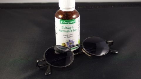 Eine Flasche Schwarzkümmelöl liegt neben einer Sonnenbrille auf einem schwarzen Untergrund, passend zum Thema Sonnenallergie.