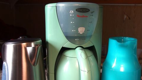 Eine grüne Kaffeemaschine steht in der Mitte zwischen einem silbernen Kessel und einer blauen Kanne auf einer Küche.