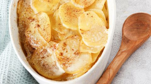 Ein Auflauf mit sahnigen Kartoffelscheiben ist warm und goldbraun gebacken, umgeben von einem hellen Tuch und einem Holzlöffel.