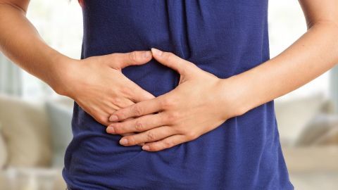 Eine Person hält ihren schmerzenden Bauch im blauen Shirt in einem gut beleuchteten Raum, was Verdauungsprobleme symbolisiert.
