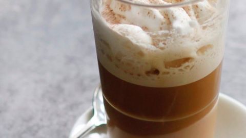 Ein Glas Frappe mit Schaumkrone steht auf einem Unterteller, ein Löffel daneben, in einem neutralen Umfeld.
