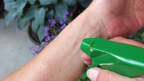 Eine Hand sprüht mit einer grünen Dusche auf den Unterarm, um sich im Garten zu erfrischen, umgeben von Pflanzen.