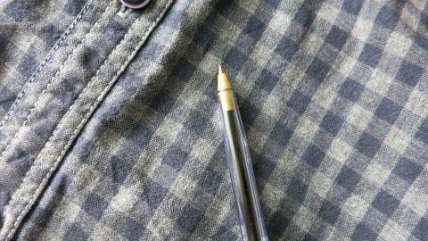 Ein Hemd mit einem Kugelschreiber daneben zeigt einen Fleck, der von Tinte auf dem karierten Stoff stammt.