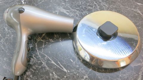 Ein Föhn liegt neben einem glänzenden Topfdeckel auf einer dunklen Arbeitsfläche, bereit, den Deckel zu lösen.