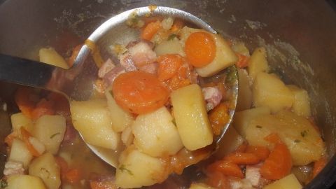 Ein Löffel hebt einen herzhaften Eintopf mit Kartoffeln und Karotten aus einem großen Topf in einer modernen Küche.