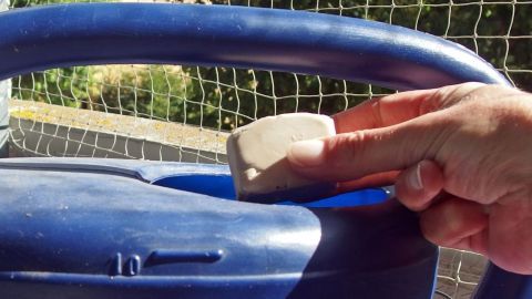 Eine Hand streicht über den blauen Griff eines Blumendüngers, während im Hintergrund ein grünes Gartenumfeld sichtbar ist.