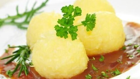 Runde Knödel liegen auf einer schmackhaften Bratensoße, garniert mit frischen Kräutern, auf einem weißen Teller.