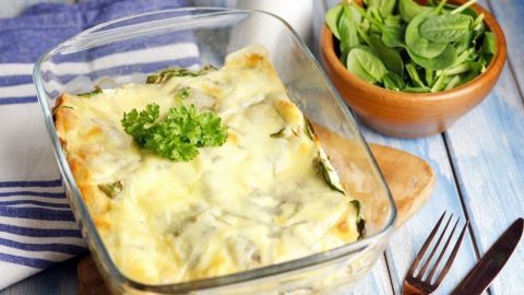 Eine goldbraune Spinat-Lasagne in einer Glasform steht auf einem Holzbrett, umgeben von einer Schüssel frischem Spinat.