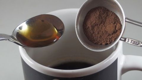 Eine Kaffeetasse enthält dunklen Kaffee, während ein Löffel Honig und ein Messlöffel Kakao darüber gehalten werden.
