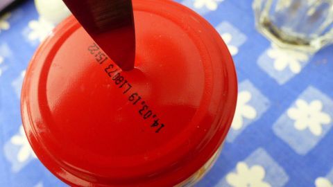 Ein roter Deckel wird mit einem L&ouml;ffel ge&ouml;ffnet, w&auml;hrend er auf einem blauen Tisch mit wei&szlig;en Blumenmuster liegt.