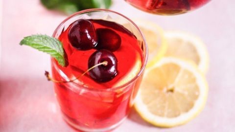 Ein Krug mit Kirschbowle wird über ein Glas mit roten Kirschen und Minze gegossen. Zitronenscheiben liegen daneben.