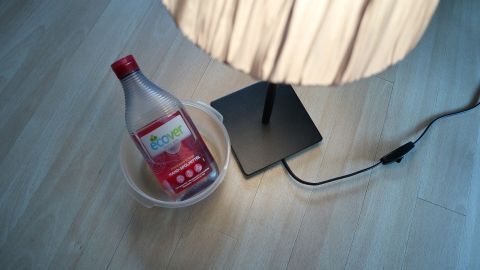 Eine Flasche mit einer roten Flüssigkeit steht in einem Schälchen neben einer Lampe auf einem Holzfußboden.