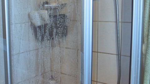 Eine Glas-Duschkabine wird mit Wasser besprüht und hat Schaumrückstände, steht in einem gefliesten Badezimmer.