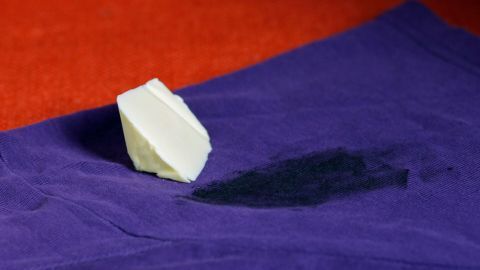 Autoschmierflecken können mithilfe einer Butter-Vorbehandlung gut entfernt werden. Ein Stück Butter liegt auf einem lila Tuch mit einem Flecken von Autoschmiere, während es zur Entfernung des Flecks verwendet wird.