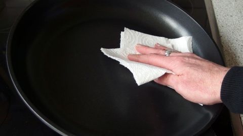 Die Pfanne mit Küchenpapier vom Fett befreien (Hausmüll) und dann erst waschen. Eine Hand wischt mit Küchenpapier altes Fett aus einer Pfanne, während der Hintergrund eine Küche zeigt.