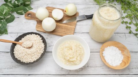 Du magst keine harten Zwiebeln im Salat? Dann probiere mal diesen einfachen Tipp für weichere Zwiebeln aus! Zwiebeln liegen auf einem Holzbrett, während ein Salat mit Salz und Reisbrei in Schalen vorbereitet wird.