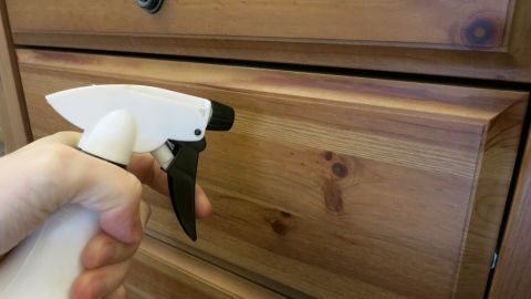 Eine Hand hält einen Sprüher, der Weichspüler auf eine glänzende Holzoberfläche eines Möbels sprüht.