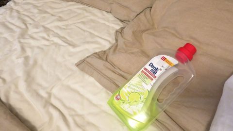 Ein super Trick für Spätaufsteher: Tupft euch etwas flüssiges Reinigungsmittel hinter die Ohren. Es wird so aussehen, als ob ihr den ganzen Tag saubergemacht hättet. Auf einem Bett liegt ein schmutziges, zerknittertes Bettlaken, neben dem eine Flasche Spülmittel positioniert ist.