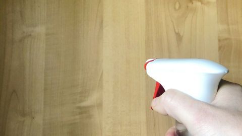 Eine Hand hält eine Sprühflasche mit Glasreiniger, der auf eine Holzoberfläche gerichtet ist, um Fettflecken zu entfernen.