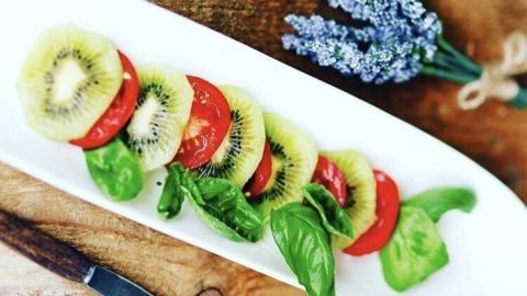 Ein bunter Kiwi-Tomatensalat liegt auf einem weißen Teller, umgeben von frischem Basilikum und Lavendel.