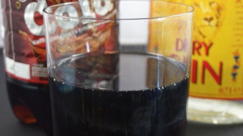 Ein Glas mit dunkler Flüssigkeit steht im Vordergrund, während eine Flasche Cola und eine Flasche Gin im Hintergrund sichtbar sind.