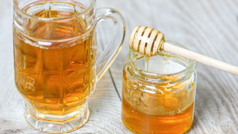 Eine Tasse mit goldenem Tee steht neben einem Glas Honig, das mit einem Holzlöffel bedeckt ist, auf einer Holzoberfläche.