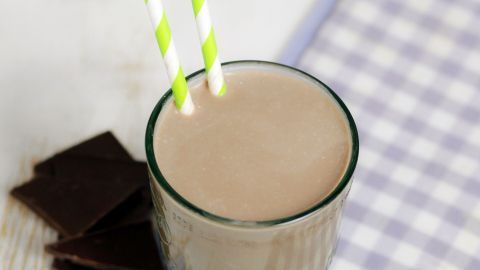 Ein Glas Schokoladenmilchshake steht auf einem karierten Tuch und hat zwei grüne Streifenhälmchen. Schokoladenstückchen liegen daneben.