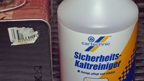 Mit Kaltreiniger, den man z. B. zum Reinigen von Automotoren o. ä. in der Sprühflasche bekommt, kann man Aufkleber entfernen. Ein Kaltreiniger steht neben einem Objekt mit einem Aufkleber, der entfernt werden soll, auf einem rauen Untergrund.