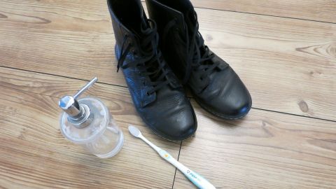 Schwarze Lederschuhe liegen auf einem Holzboden, neben ihnen eine Zahnbürste und ein Seifenspender für die Reinigung.