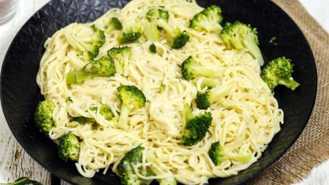 Eine Pfanne mit Spaghetti und Broccoli zeigt das Gericht, umgeben von Olivenöl und rohen Spaghetti auf einem rustikalen Tisch.