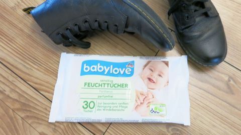 Schwarze Lederschuhe liegen neben einer Packung Babyfeuchttücher auf einem Holzboden, bereit zum Putzen.