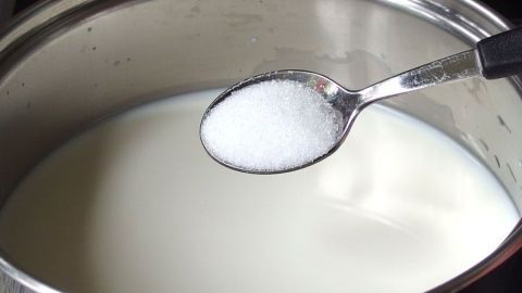 Im Topf befindet sich Milch, während ein Löffel mit Zucker darüber gehalten wird; die Szene zeigt eine Kochumgebung.