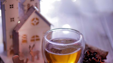 Ein Glas warmer Met steht auf einem Holztisch, umgeben von Zimtstangen, einem Tannenzapfen und einer festlichen Kulisse.