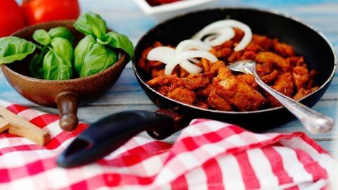 In einer Pfanne brutzelt das Tiefkühl-Gyros, umgeben von frischen Kräutern und Tomaten auf einem karierten Tuch.