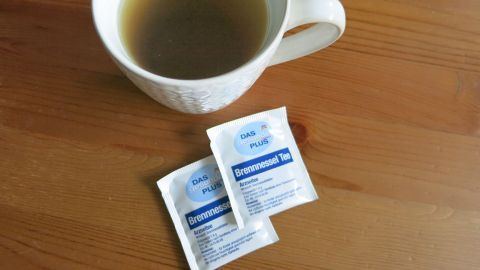 Eine Tasse Brennnesseltee steht neben zwei Packungen Teebeuteln auf einem Holztisch.